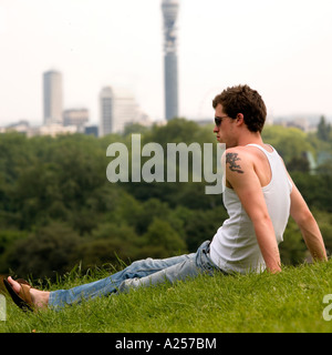 Junger Mann sitzt auf Primrose Hill mit der Londoner Skyline im Hintergrund im Sommer Stockfoto