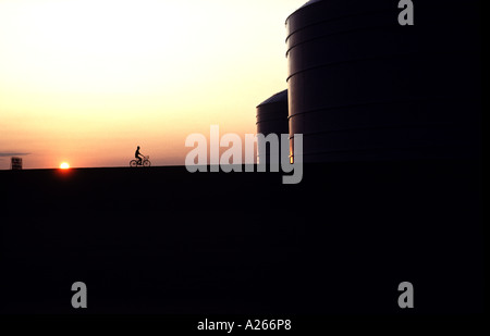 Silhouette eines Mannes, der sein Fahrrad um Ölfeld Panzer bei Sonnenaufgang. Stockfoto