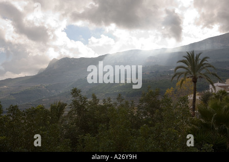 Guadalest Valley mit Sonnenstrahlen durchbrechen Wolken am fernen Berge und Palme im Vordergrund Stockfoto