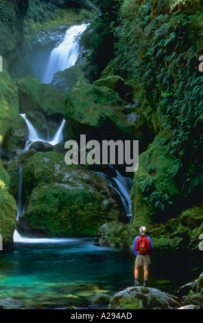 Wanderer mit Rucksack auf einem Felsen am Fuße eines Wasserfalls, umgeben von Felsen, Moos bedeckt Stockfoto