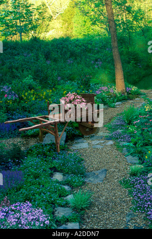 Mit Blumen gefüllte antike Schubkarre auf einem Schotterweg in einem idyllischen schattigen Garten entlang des Pathway, Missouri, USA Stockfoto