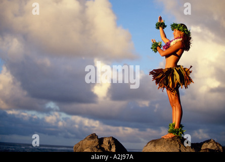 Native Hawaiian Frau trägt ein Rasen Rock Farn Blatt Armbänder und bunten Blumen Leis steht auf einem Felsen mit Blick auf den Ozean Stockfoto