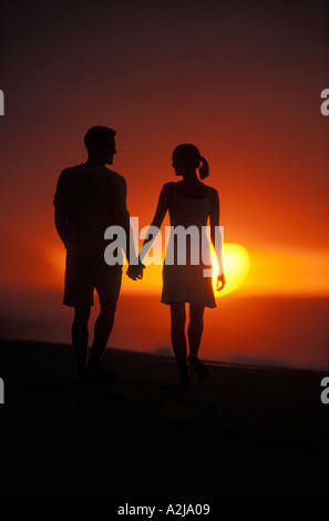 Lässig gekleidet, barfuß paar Silhouette durch die Einstellung Sonne Spaziergang hand in hand an einem Strand entlang Stockfoto