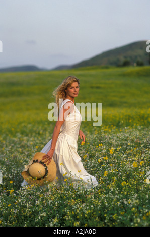 Blonde Frau trägt eine weiße Sonnenkleid mit einem Strohhut geht in einem Feld von kleinen weißen und gelben Wildblumen Stockfoto
