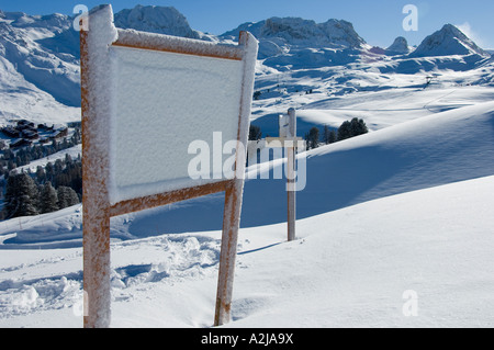 Schnee bedeckte Schild auf einem weißen Schneefeld mit Bergen und einem Alpendorf Stockfoto