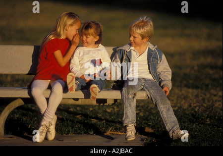 Kleines Mädchen sitzt auf einer Bank zwischen einem jungen Mädchen flüstert in ihr Ohr und ein kleiner Junge versucht, ihr eine einzelne rote Rose schenken Stockfoto