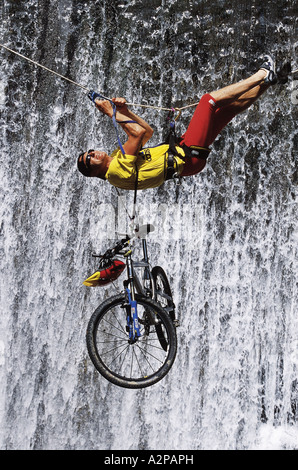 Mountainbiker mit seinem Fahrrad überqueren einer Schlucht hängen an einem Kletterseil, Wasserfall im Hintergrund Stockfoto