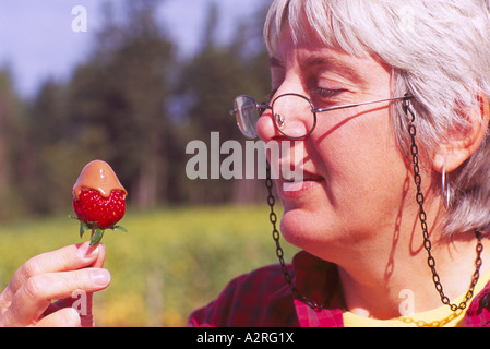 Reife behaarte grau / grau dunkelhaarige Frau mit Brille essen Schokolade getaucht Erdbeere Stockfoto