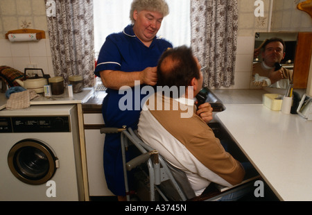 Bezirk Krankenschwester hilft die Haarbürste eines behinderten Mannes in seinem Rollstuhl bei einem Besuch in seinem Haus in London UK Stockfoto