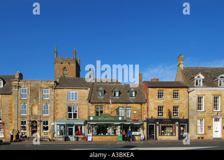 Erleben Sie den Tag des blauen Himmels der Wold in der Straßenszene der Stadt Cotswolds Market mit Kirchturm dahinter in einem Teil des Market Square Gloucestershire England Großbritannien Stockfoto
