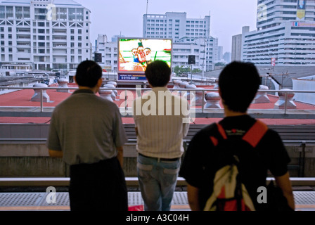 Drei Skytrain Pendler vor dem Fernseher auf einem großen Monitor auf dem Bahnsteig. Stockfoto