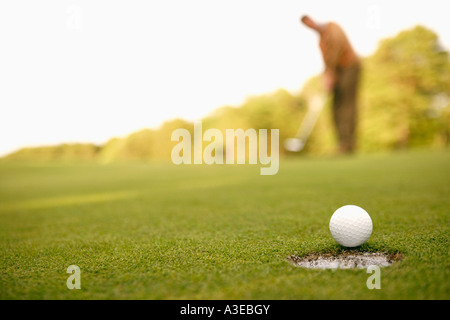 Nahaufnahme eines Golfballs am Rand eines Lochs mit einem Mann im Hintergrund Stockfoto