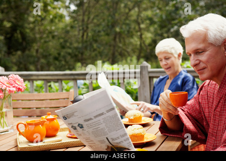 Nahaufnahme von einem reifen Mann hält eine Tasse und liest eine Zeitung mit einer älteren Frau im Hintergrund Stockfoto