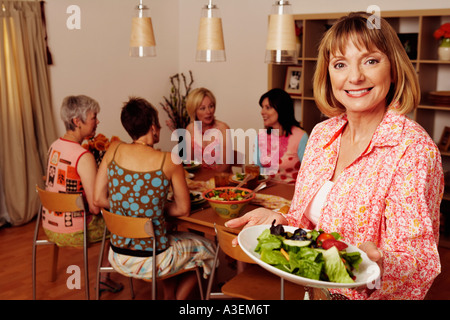 Porträt einer reifen Frau einen Teller mit Salat und ihre Freunde sitzen am Esstisch im Hintergrund halten Stockfoto