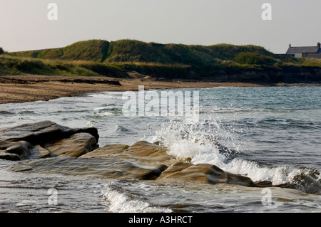 Wellen brechen sanft über Felsen und schlendern mit grasbewachsenen Sanddünen am Strandrand in der Ferne. Northumberland Großbritannien Stockfoto