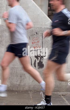 Jogger laufen vorbei an Banksy Schablone Graffiti Ratte mit Go Back To Bed-Schild. Thames Embankment, London, England Stockfoto