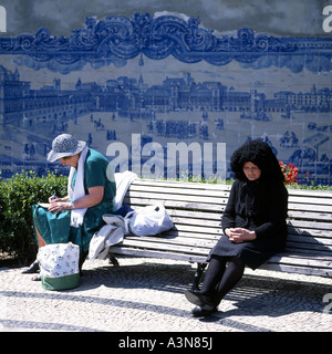 ÄLTERE PORTUGIESISCHE FRAU MIT TRADITIONELLEM SCHWARZEN KLEID UND REIFE TOURISTENKARTE, DIE AUF EINER BANK IM STADTTEIL ALFAMA IN LISSABON, PORTUGAL, EUROPA, EINE POSTKARTE SCHREIBT Stockfoto