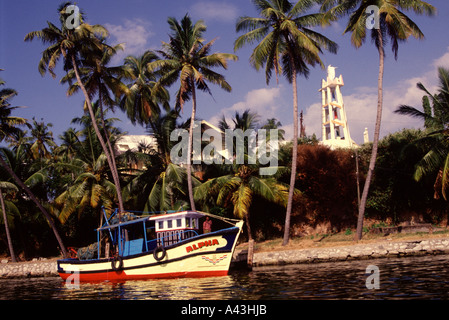 Ein Holzboot, das in einem palmengesäumten Kanal mit einem Glockenturm einer Kirche im Hintergrund in Kerala Backwaters Südindien festgemacht ist Stockfoto