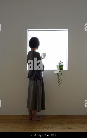 Rückansicht einer jungen Frau hält eine Kaffeetasse und stand in der Nähe eines Fensters Stockfoto