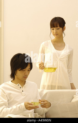 Junger Mann hält eine Tasse Tee mit einer jungen Frau hält eine Teekanne hinter ihm Stockfoto