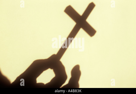 Nahaufnahme eines Schattens an einer Creme Wand im warmen Licht der kleinen Kruzifix Begriffen zwischen Daumen und Zeigefinger Stockfoto