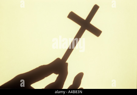 Nahaufnahme eines Schattens an einer Creme Wand im warmen Licht der kleinen Kruzifix Begriffen zwischen Daumen und Zeigefinger Stockfoto