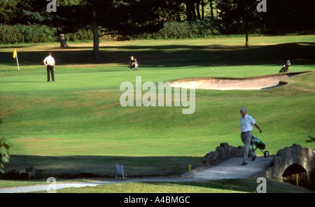 Ein älterer Mann mit Golfschläger und Trolley geht über eine Brücke, Golfplatz, wie andere Golfer im Hintergrund setzen Stockfoto