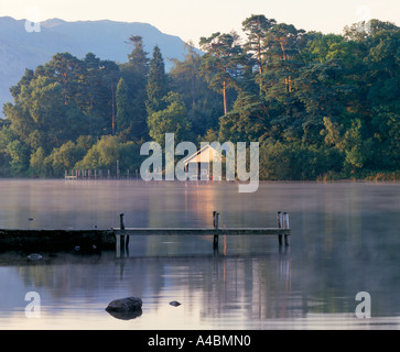 Ein sonnendurchflutetes Bootshaus umrahmt von altem Baumbestand am See Derwent im englischen Lake District mit Nebel auf den See und einem Steg liegend Stockfoto