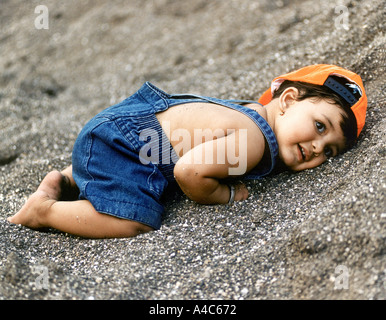 Ein kleiner Junge ein Jeans-Kleid und eine Mütze tragen liegt auf dem sand Stockfoto