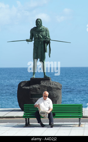 Ältere Mann lesen Zeitung Bank, beobachtet von ANATERVE eine Guanchen-Statue in Candelaria Teneriffa-Kanarische Inseln-Spanien Stockfoto