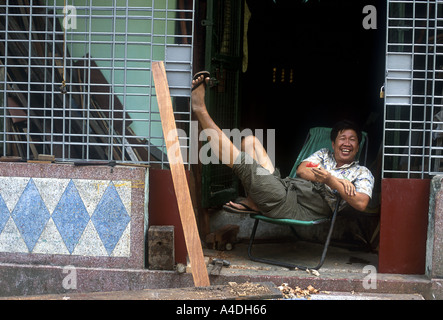 Ein Mann zum Entspannen in einem Stuhl mit den Füßen, in der Tür zu seinem Laden. Yangon, Rangun, Myanmar, Birma Stockfoto