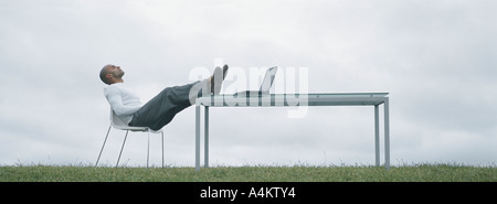 Mann sitzt im Stuhl im Freien mit Füßen auf Tisch und Laptop am Tisch, vor bedecktem Himmel Stockfoto