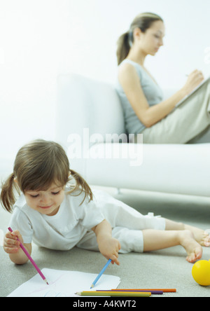 Kleines Mädchen liegend am Boden Färbung, junge Frau sitzt hinter ihr auf Couch mit Knien verfassen Stockfoto