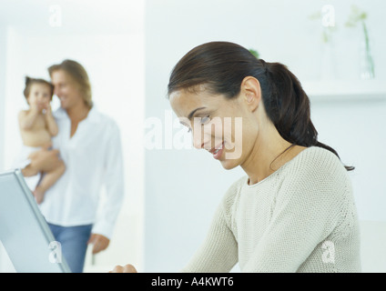 Junge Frau blickte auf Laptop, jungen Mann, der kleine Mädchen im Hintergrund hält Stockfoto