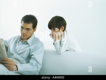 Mann sitzt auf dem Sofa lesen Zeitung, junge stand hinter Sofa mit Blick auf die Schulter des Mannes Stockfoto