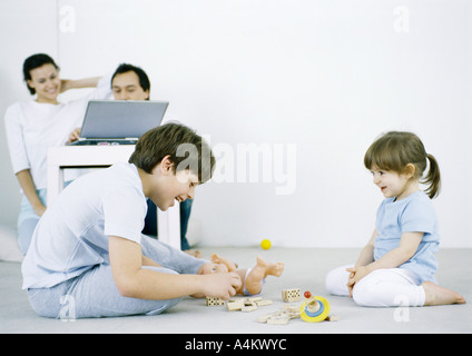 Jungen und Mädchen sitzen auf dem Boden spielen, Mann und Frau mit Laptop im Hintergrund Stockfoto