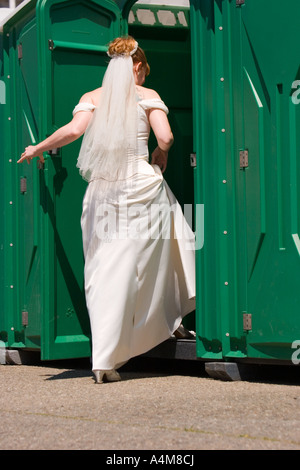 Eine Braut im weißen Kleid und Schleier betritt ein Port-a-Töpfchen. Stockfoto