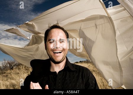 Attraktiver Mann in seinem 20 s 30 s lacht außerhalb in einem Feld hohem Gras an einem windigen sonnigen bewölkten Tag mit weißen Laken bläst. Stockfoto