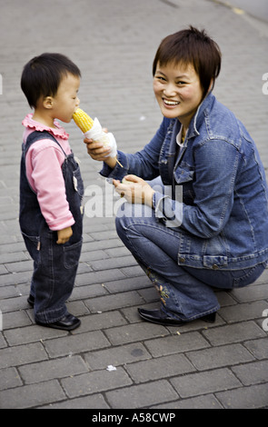 CHINA SHANGHAI modernen jungen chinesischen Mutter in Jeans gekleidet hilft Tochter Kleinkind einen Snack von geröstetem Mais zu essen Stockfoto