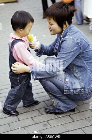 CHINA SHANGHAI modernen jungen chinesischen Mutter in Jeans gekleidet hilft Tochter Kleinkind einen Snack von geröstetem Mais zu essen Stockfoto