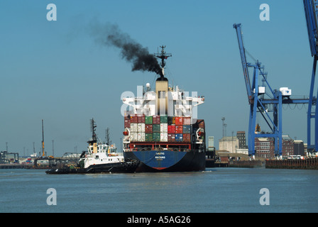 Frachtschiff mit schwarzem Rauch-Container-Frachtschiff gestapelte Ladung von Frachtcontainern Schlepper schiebt zu Kränen, die im Felixstowe-Hafen Suffolk UK ankommen Stockfoto