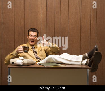 Caucasion Mitte Erwachsenen Retro-Geschäftsmann, sitzen mit Füßen gestützt auf Schreibtisch Kaffeetrinken geben einen Daumen nach oben Stockfoto