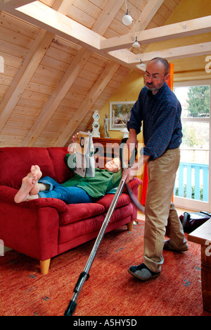 Herr PR-Mann Staubsaugen, Staubsaugen unter eine Frau liegt auf dem Sofa Lesen einer Zeitung Stockfoto