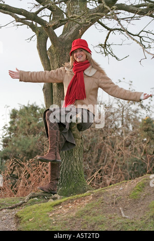 Junge Frau trägt eine rote Mütze und Schal. Blondes Haar. Natürlich. Auf dem Ast eines Baumes sitzt. New Forest, Hampshire. VEREINIGTES KÖNIGREICH. Stockfoto