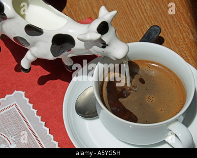 gießt Milch aus einem Milchkännchen in Form einer Kuh in eine Tasse Kaffee am roten Tisch mit einem Deckchen topview Stockfoto