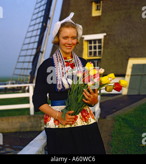 Niederländische Mädchen in Tracht Holding Haufen gelbe und rote Tulpen mit Windmühle hinter ihrem Niederlande Stockfoto