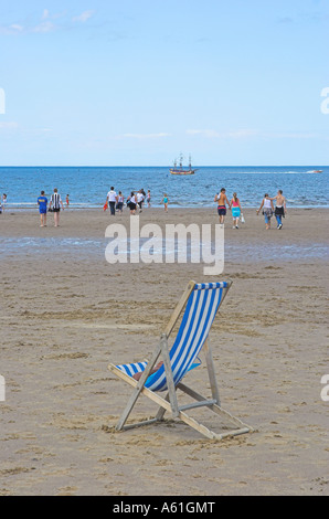 Einem einzigen blau-weiß gestreiften Liegestuhl auf Whitby Strand mit Sommerhimmel und Menschen spielen in der Ferne Küste Stockfoto