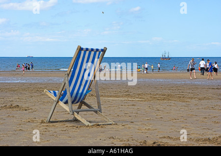 Einem einzigen blau-weiß gestreiften Liegestuhl auf Whitby Strand an einem Sommertag mit Menschen spielen am fernen Ufer Stockfoto