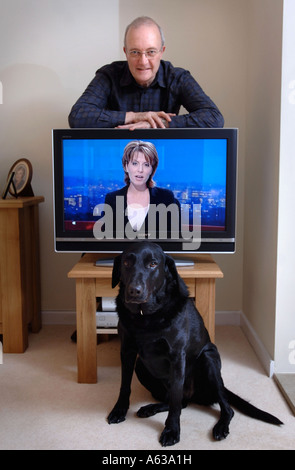 EIN APPLYING MANN LEHNT SICH AN EINEN FERNSEHER, WÄHREND EIN PET SCHWARZ LABRADOR AUF BRITISCHEN BODEN SITZT Stockfoto