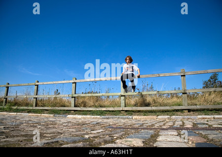 Langhaarige junge sitzt auf einem hölzernen Zaun durch einen gepflasterten Weg mit einem großen blauen Himmel und ein Feld der Trockenrasen Stockfoto